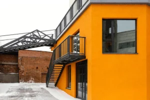 A vibrant orange building with black trim and stairs, connected by a covered walkway to a brick structure, set against a snowy backdrop.