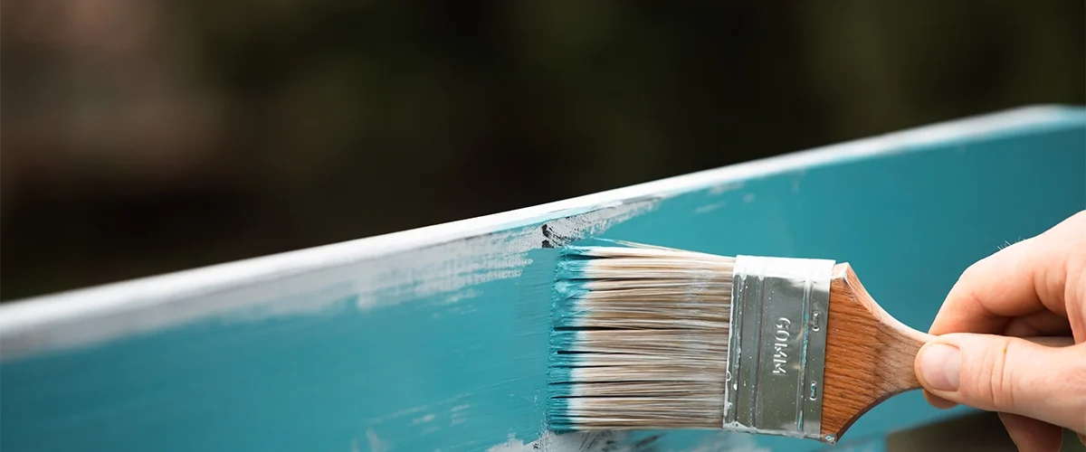 Applying teal paint to a wooden surface with a paintbrush.