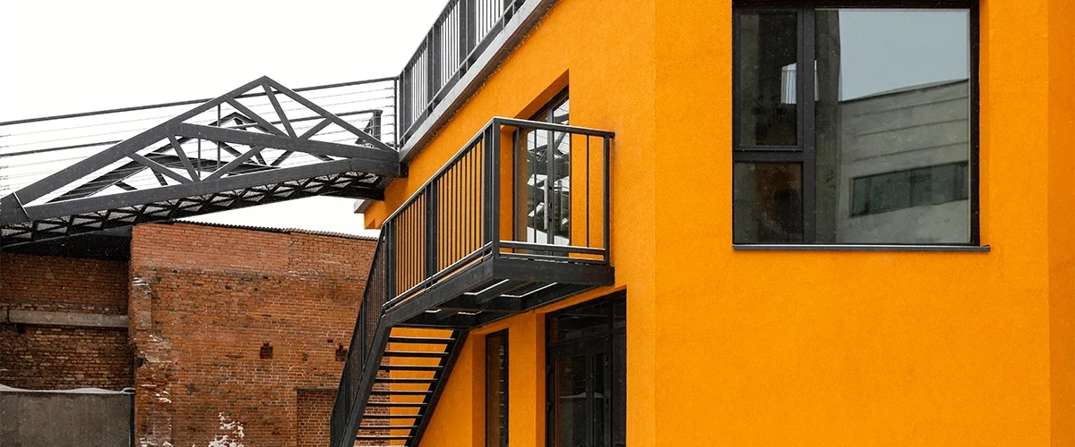 A vibrant orange building with black trim and stairs, connected by a covered walkway to a brick structure, set against a snowy backdrop.