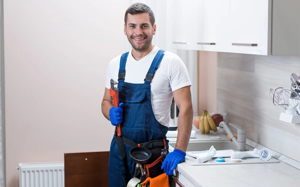 A smiling plumber holding a wrench and standing in a kitchen.